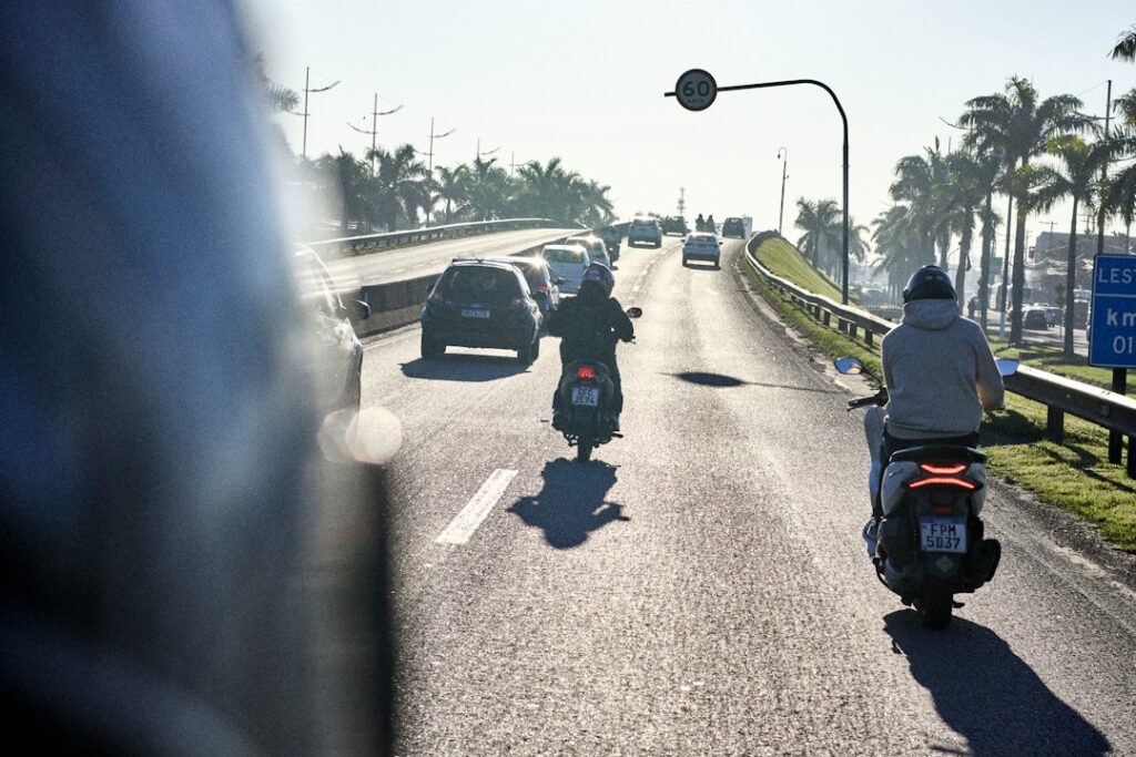 Two motorcyclists riding on a highway with cars in the distance under bright light